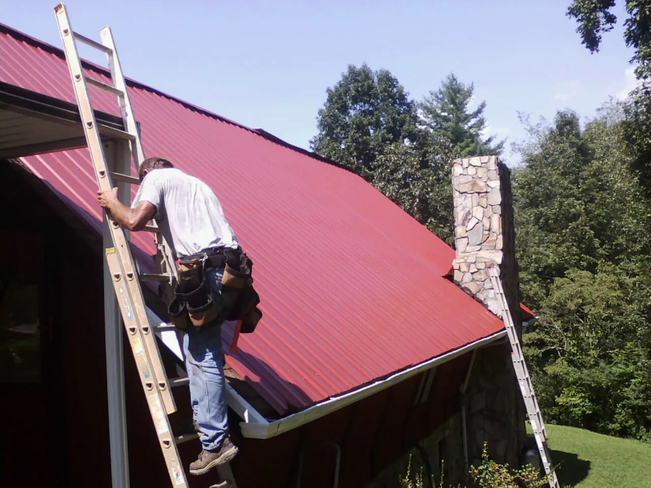 Roofer on a ladder beside a red metal roof and stone chimney — Soffit & Fascia Repair by Roofing Brothers in Burley